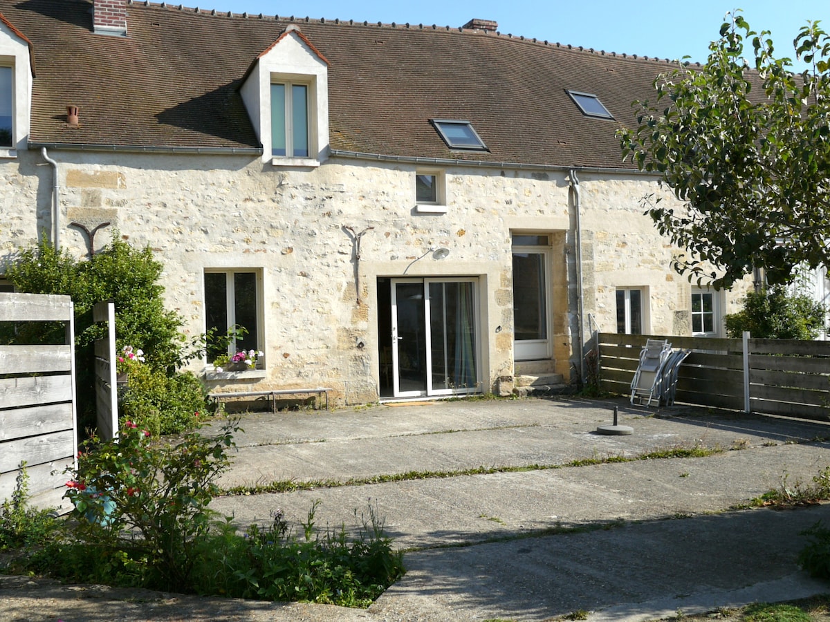 The exterior of a renovated farmhouse presents a charming stone facade, featuring multiple windows and a sliding glass door leading to a private outdoor area. A spacious courtyard is visible, surrounded by greenery and garden features, offering a serene environment for relaxation.