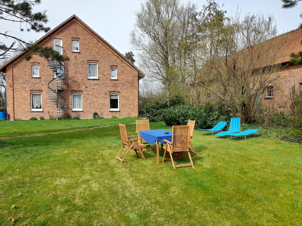 A spacious outdoor area is visible, featuring a wooden table set with chairs on a green lawn. Two lounge chairs are positioned nearby. In the background, a two-story brick building is seen, alongside another structure, with a spiral staircase visible on one side.