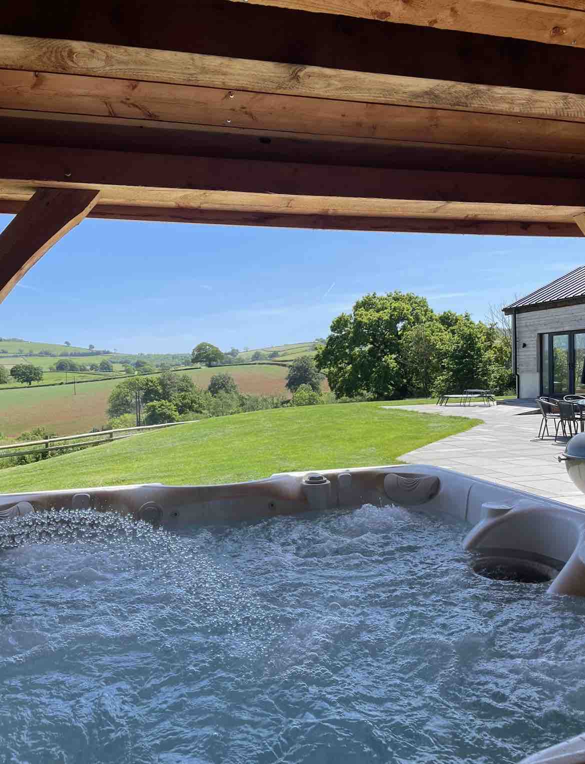 A private hot tub is situated under a wooden pavilion, with bubbling water visible. Expansive green fields and trees are showcased in the background under a clear blue sky, creating a serene outdoor setting. The surrounding patio is proportionately spacious.