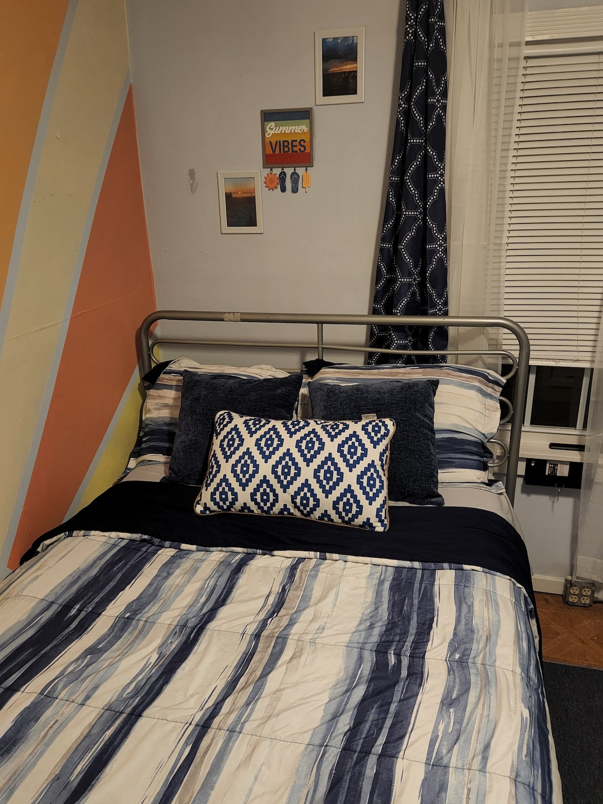 A comfortable sleeping area features a metal bed frame with striped bedding in blue and white tones. Decorative pillows add texture to the bed, while a colorful accent wall and framed artwork contribute to the room's ambiance. Natural light filters through a partially open window.