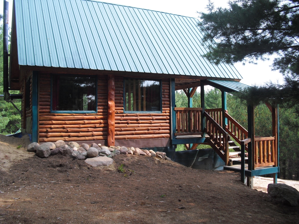 The exterior of a cabin is framed by tall pine trees, featuring a rustic log structure with a blue metal roof. A wooden staircase leads to a welcoming entryway, complemented by a natural stone border along the ground.