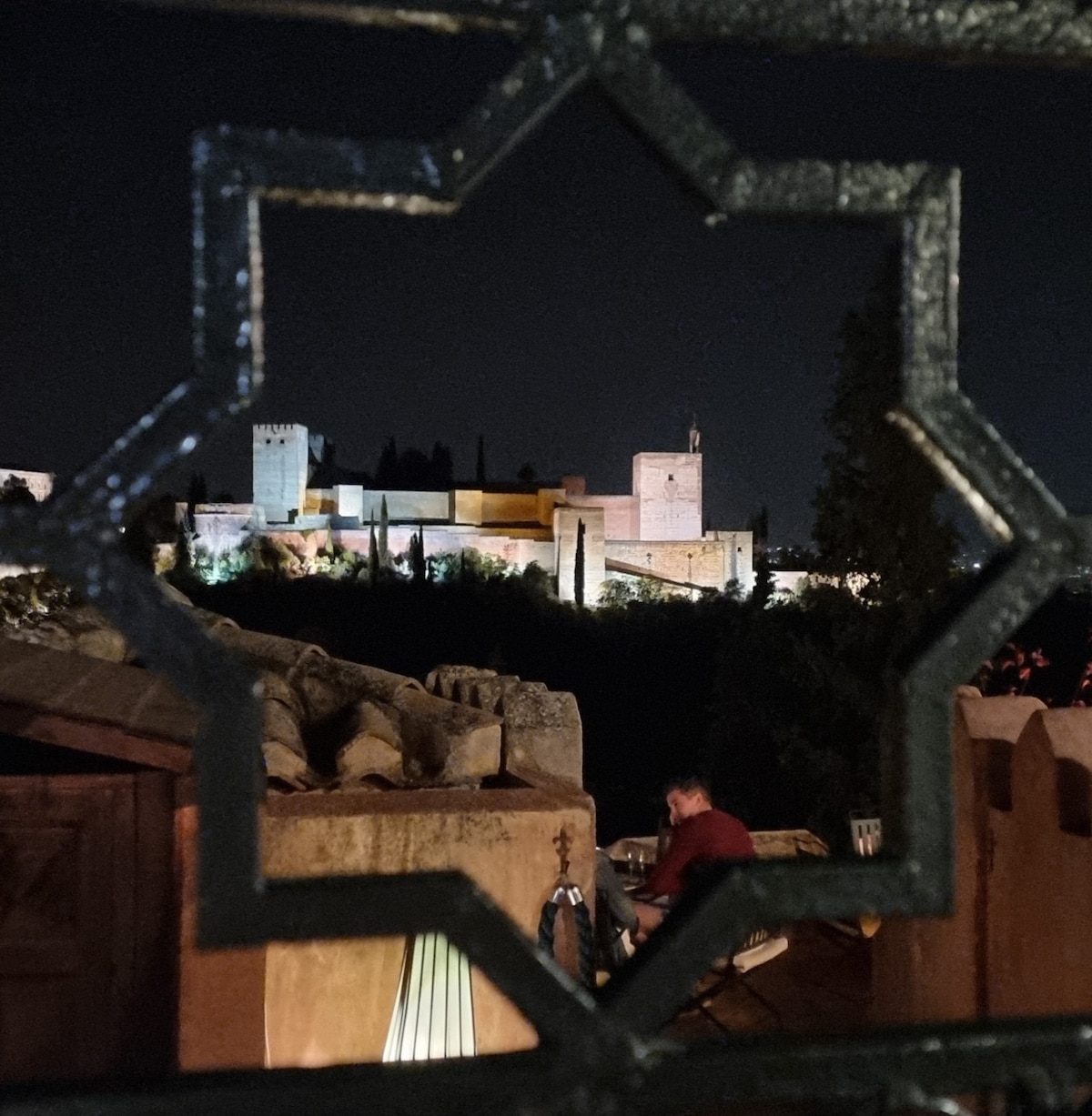 A nighttime view of a historical structure is framed by an ornamental iron window. The illuminated buildings showcase a blend of textures and colors against the dark sky, surrounded by trees that create a serene backdrop.