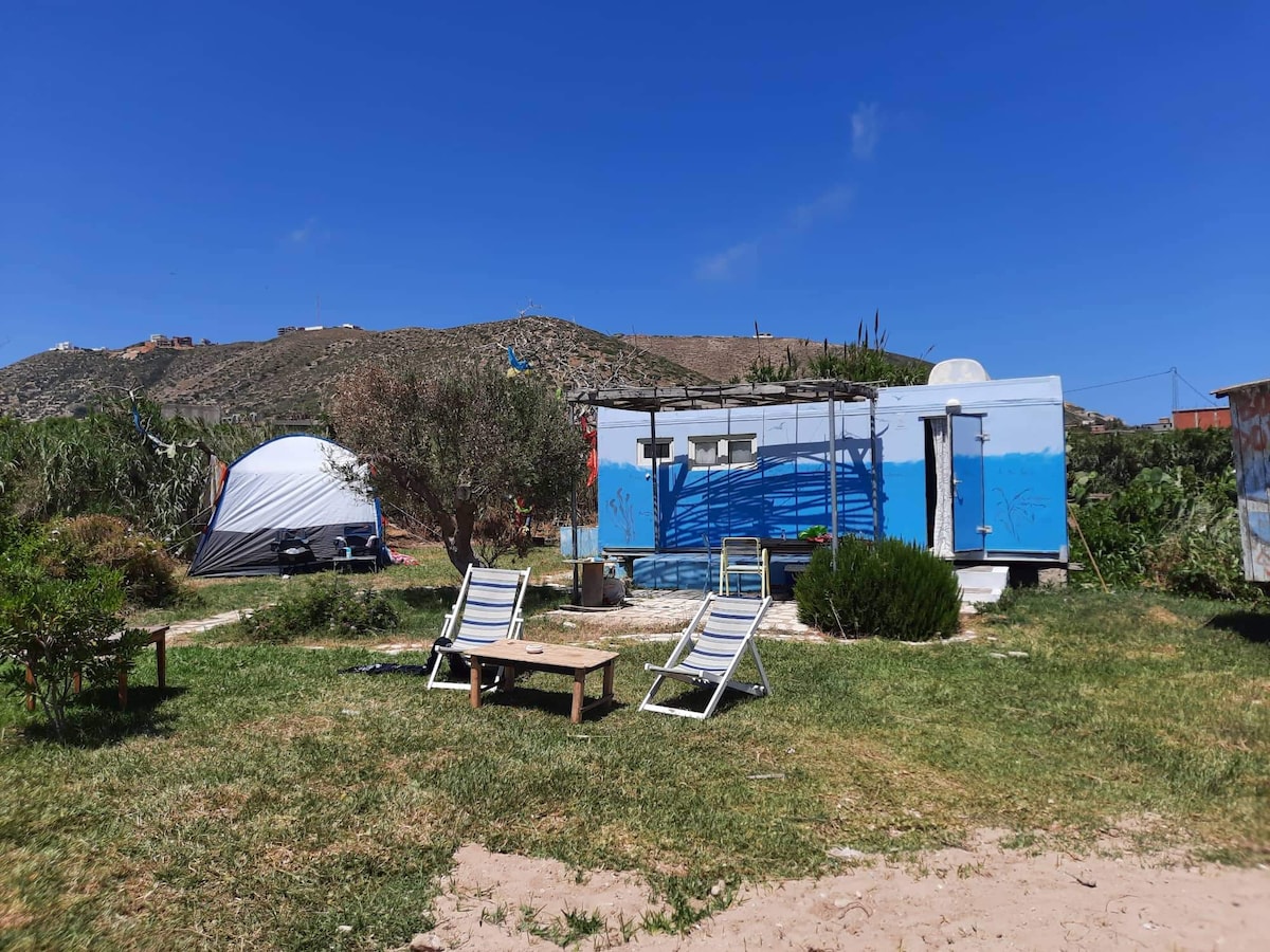 A caravan set in a grassy area is surrounded by shrubs and hills. Two lounge chairs are positioned on the grass in front of the caravan. A tent can be seen in the background, and the sky is clear with a few clouds.