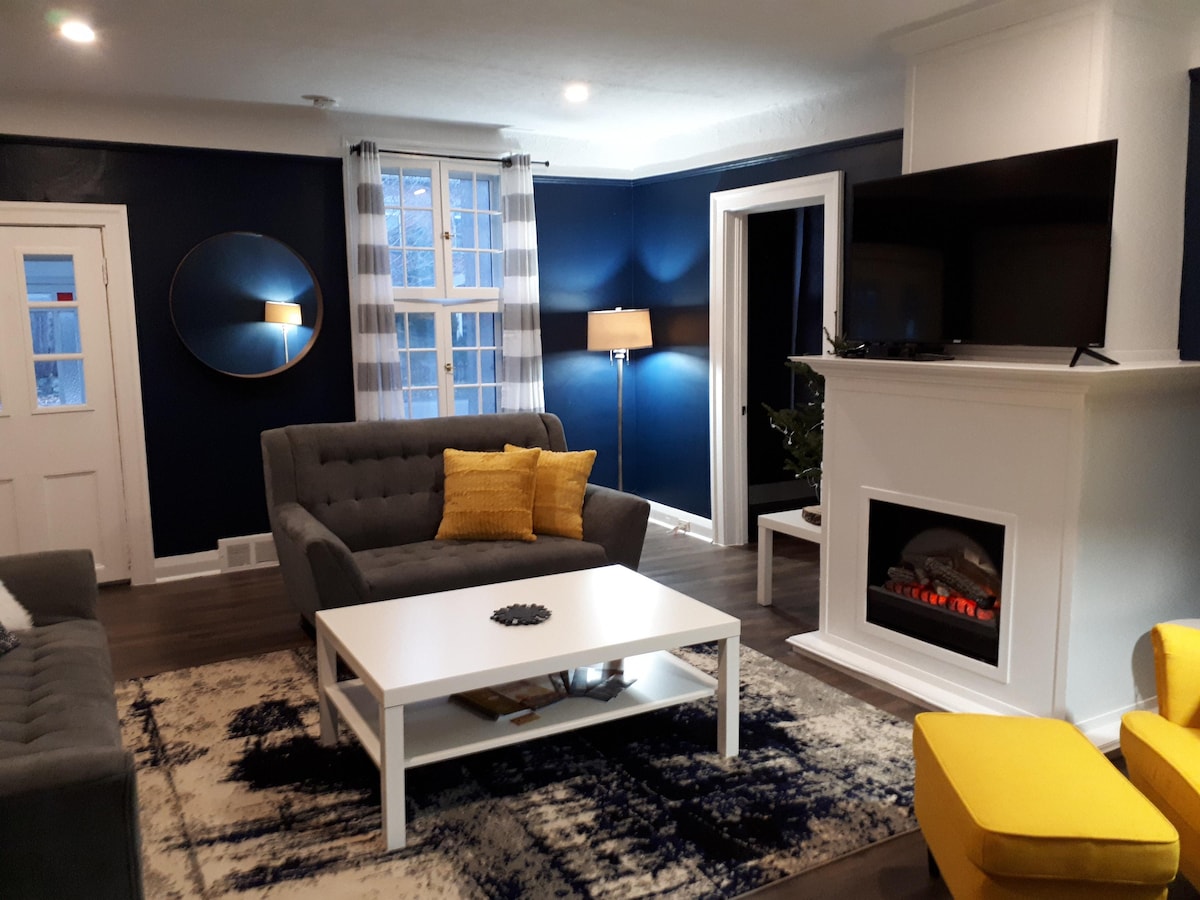 The living room features a modern aesthetic with blue walls and natural light flowing through large windows. Comfortable seating options include a gray sofa and a bright yellow armchair. A decorative fireplace is positioned below a wall-mounted television, and a coffee table rests on a contemporary area rug.