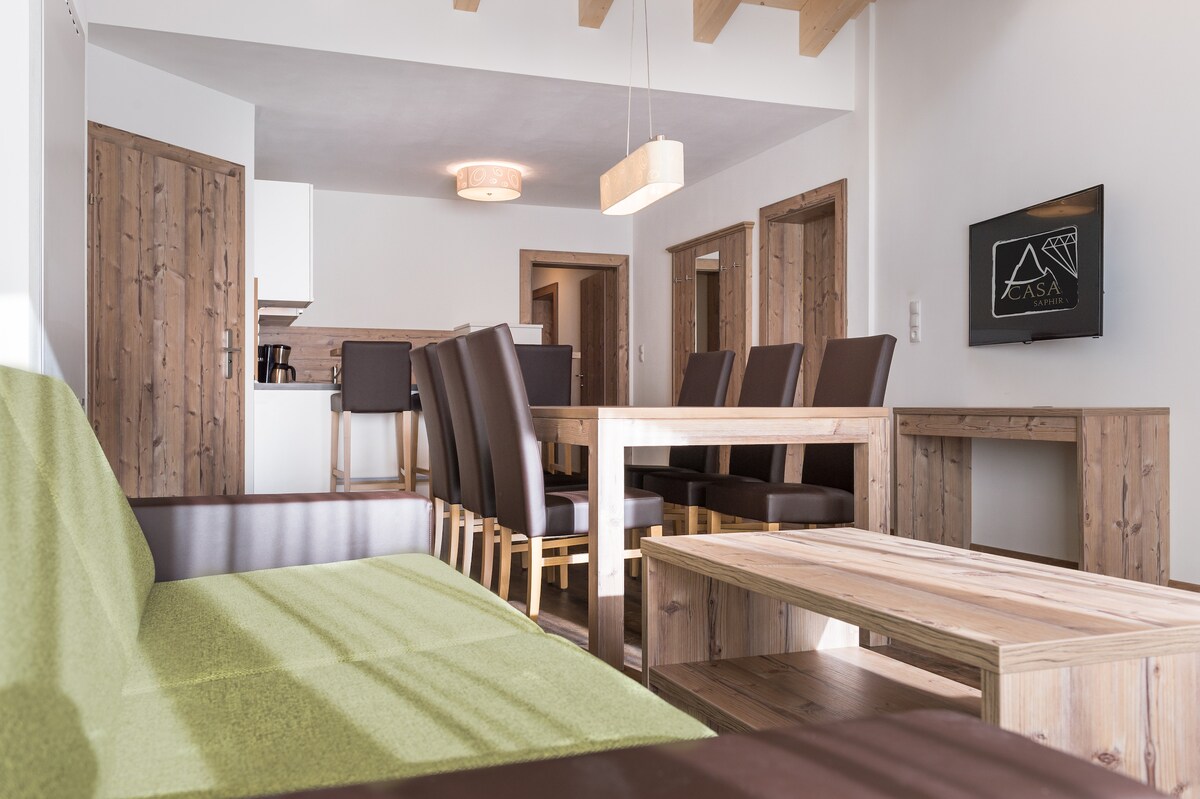 A spacious living and dining area is displayed, featuring a green sofa and a wooden coffee table. A large dining table surrounded by brown chairs is visible, along with a modern light fixture above. A flat-screen TV is mounted on the wall, and multiple doorways lead to other rooms.