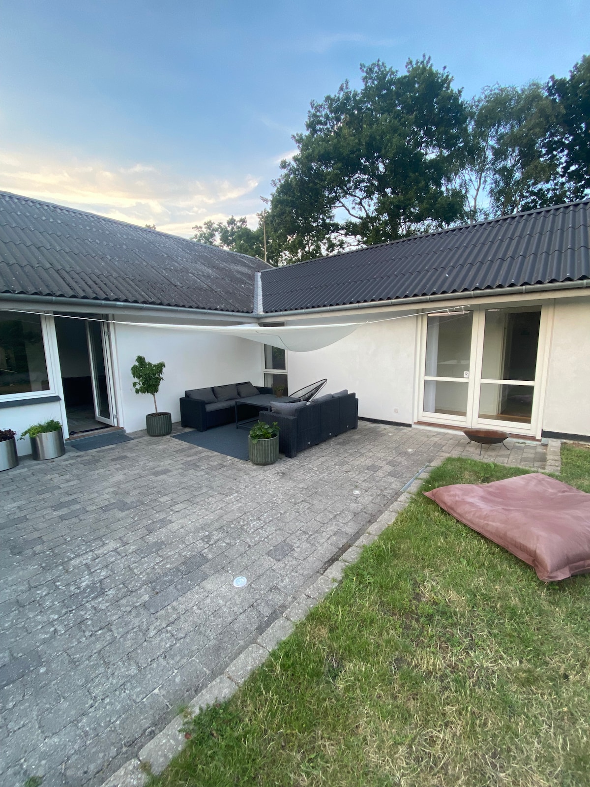 An outdoor seating area is present, featuring a modern sectional sofa surrounded by potted plants. A comfortable bean bag is positioned on the lawn. A paved pathway leads to large glass doors, inviting in natural light, with a backdrop of greenery from nearby trees.