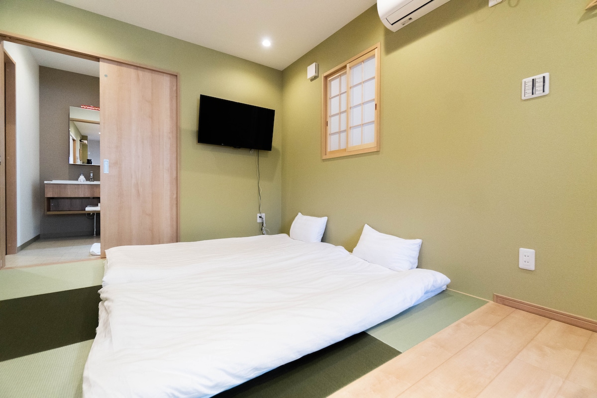 A serene sleeping area is presented with a spacious tatami mat, accommodating two soft bedding arrangements. Light green walls and wooden flooring contribute to the calming ambiance, while a wall-mounted TV and subtle air conditioning unit are visible in the background.