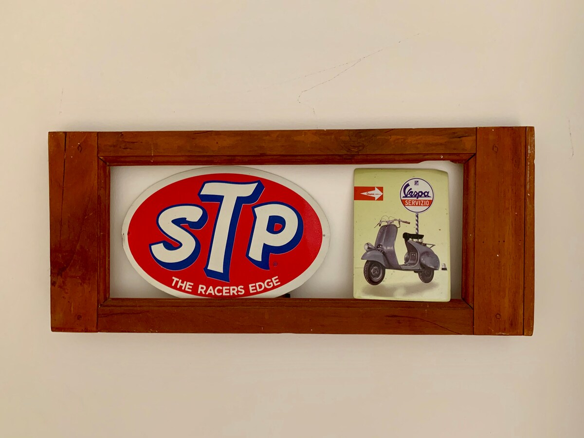 A wooden framed display features a vintage STP sign alongside a small metal sign depicting a classic Vespa scooter. The bright colors of the signs bring a retro vibe, highlighting the charm of the decor.