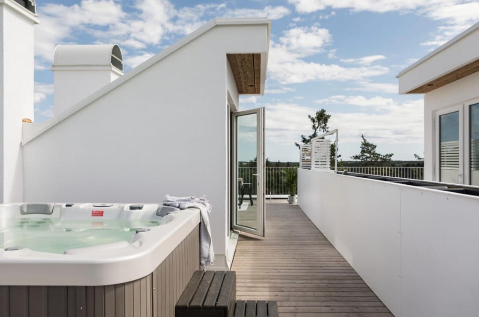 A spacious roof terrace features a jacuzzi, with a light-colored wooden deck extending towards an open door. A soft towel is draped over a dark bench, and greenery is visible in the distance, providing a sense of tranquility.