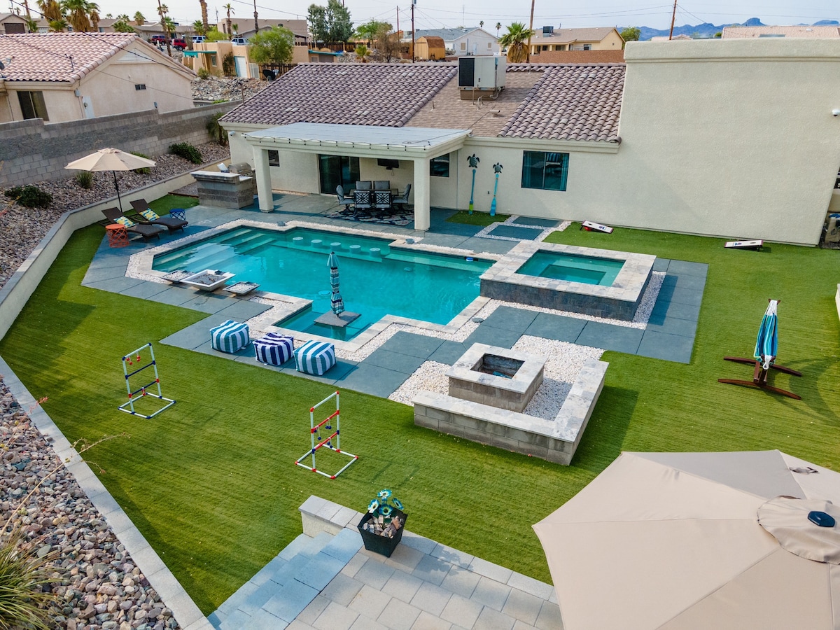 An aerial view of a private backyard features a clear blue pool surrounded by a spacious stone patio. Sun loungers are set near the water, with umbrellas providing shade. A fire pit is positioned at one corner, and a well-maintained lawn offers ample green space.