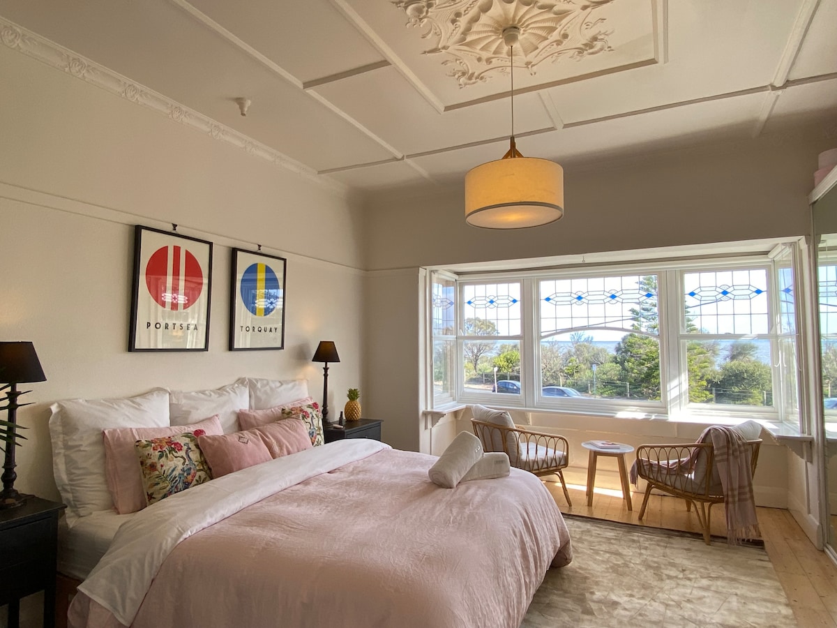 A spacious bedroom features a comfortable King bed adorned with soft linens and decorative pillows. Natural light filters through a large bay window, offering views of the beach. Two rattan chairs and a small table complement the room's design, providing a cozy seating area.