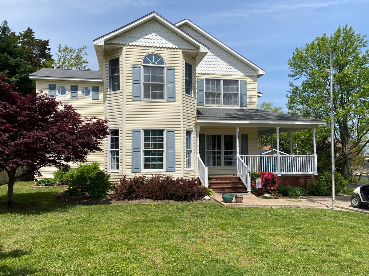 The two-story house is situated on a well-maintained lawn, characterized by landscaped gardens and a variety of shrubs. A large covered porch spans the front, framed by large windows and traditional shutters, providing ample natural light.