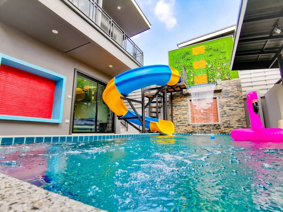 A vibrant swimming pool features a colorful water slide and a decorative stone wall. A playful pink float is visible in the water, while a gentle waterfall cascades down the stone surface. The surrounding area includes modern architectural elements and bright accent colors.