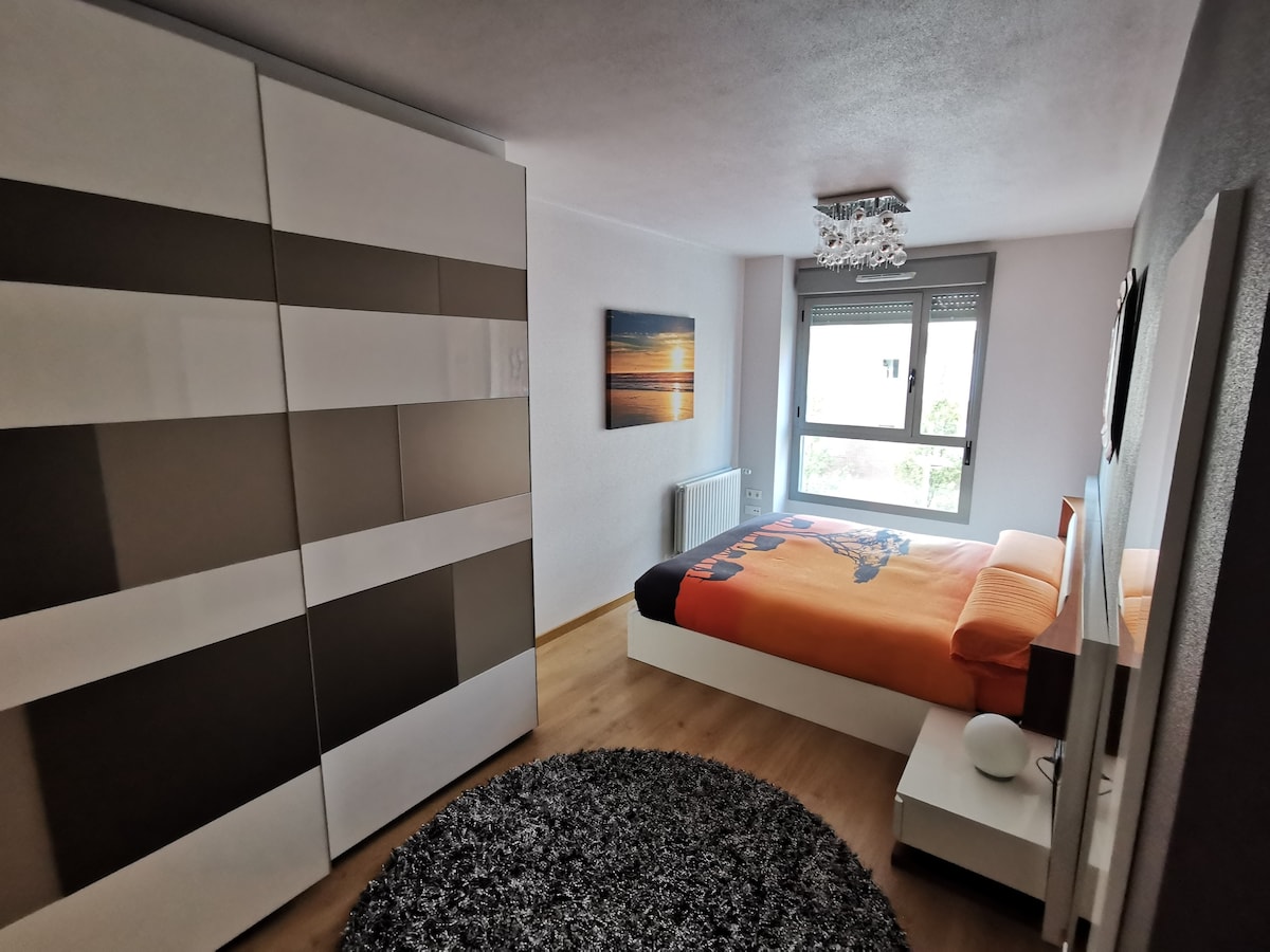 A well-furnished bedroom features a spacious bed dressed in bright orange bedding. A large wardrobe with a modern design occupies one wall, while a round rug adds texture to the wooden floor. Natural light filters in through two windows with unobstructed views.
