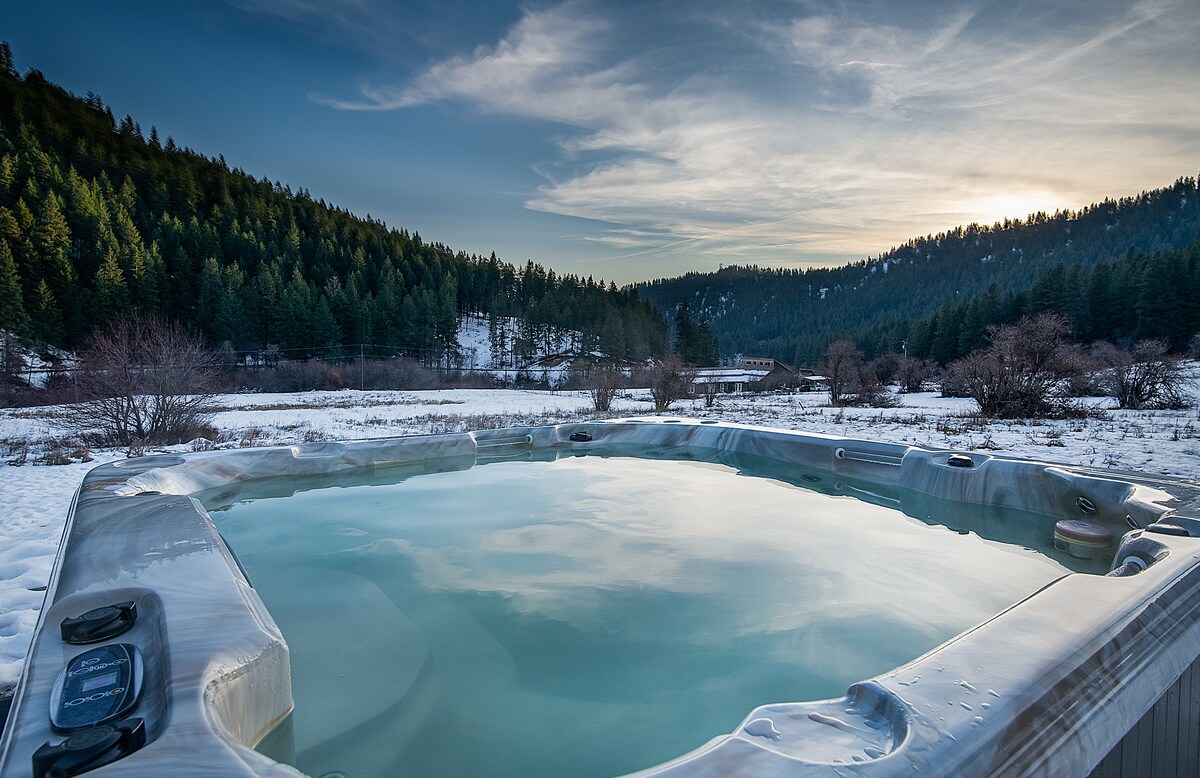 An outdoor hot tub is situated in a snow-covered landscape, surrounded by evergreen trees and rolling hills. The water's surface reflects the sky, showcasing the tranquil environment. Gentle slopes are visible in the distance, enhancing the serene setting.