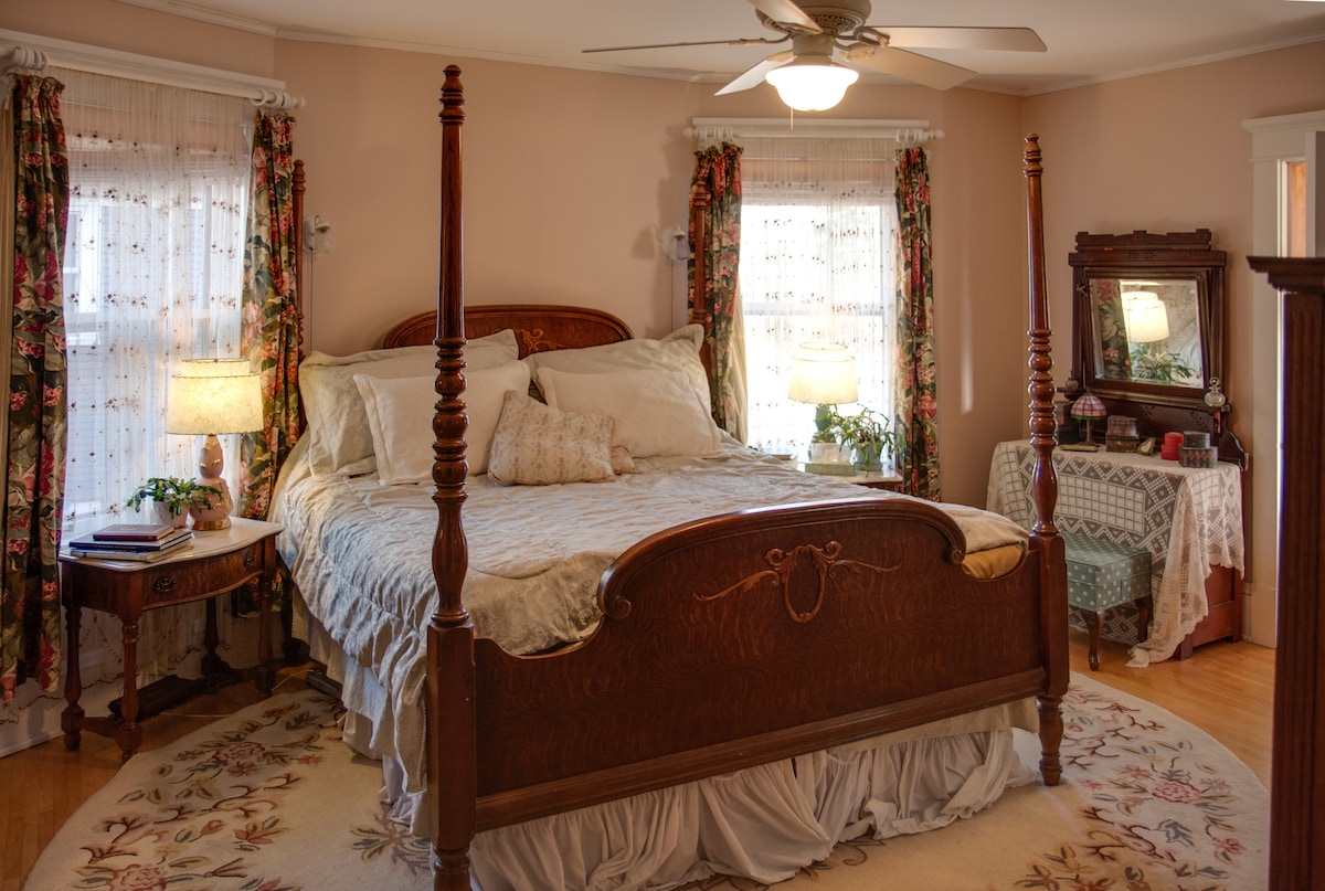 A spacious room features a king-sized four poster bed, surrounded by floral curtains and soft lighting. An elegant mirror stands on a nearby table, and a decorative rug anchors the space, enhancing the cozy ambiance with natural wooden accents.