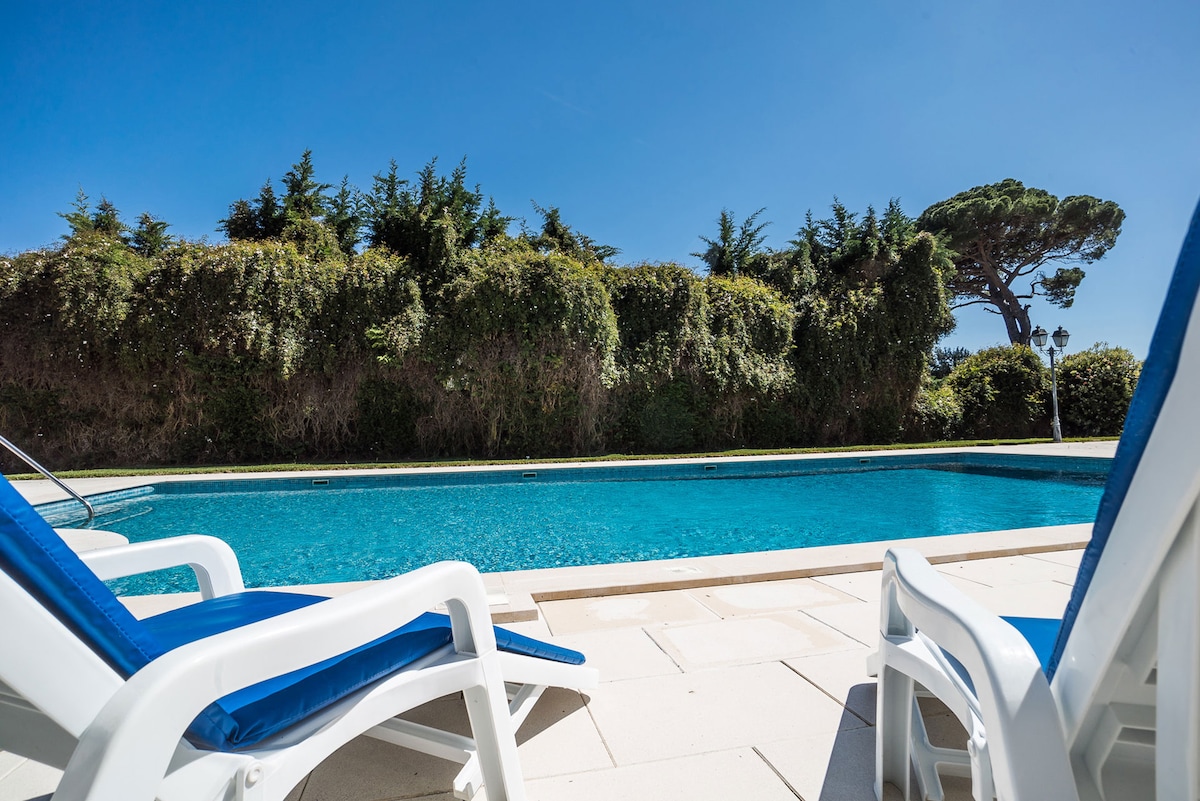 A serene swimming pool is framed by lush greenery, creating a private retreat. Two white lounge chairs with blue cushions are positioned nearby, inviting relaxation. The clear water reflects the bright blue sky, enhancing the tranquil outdoor atmosphere.