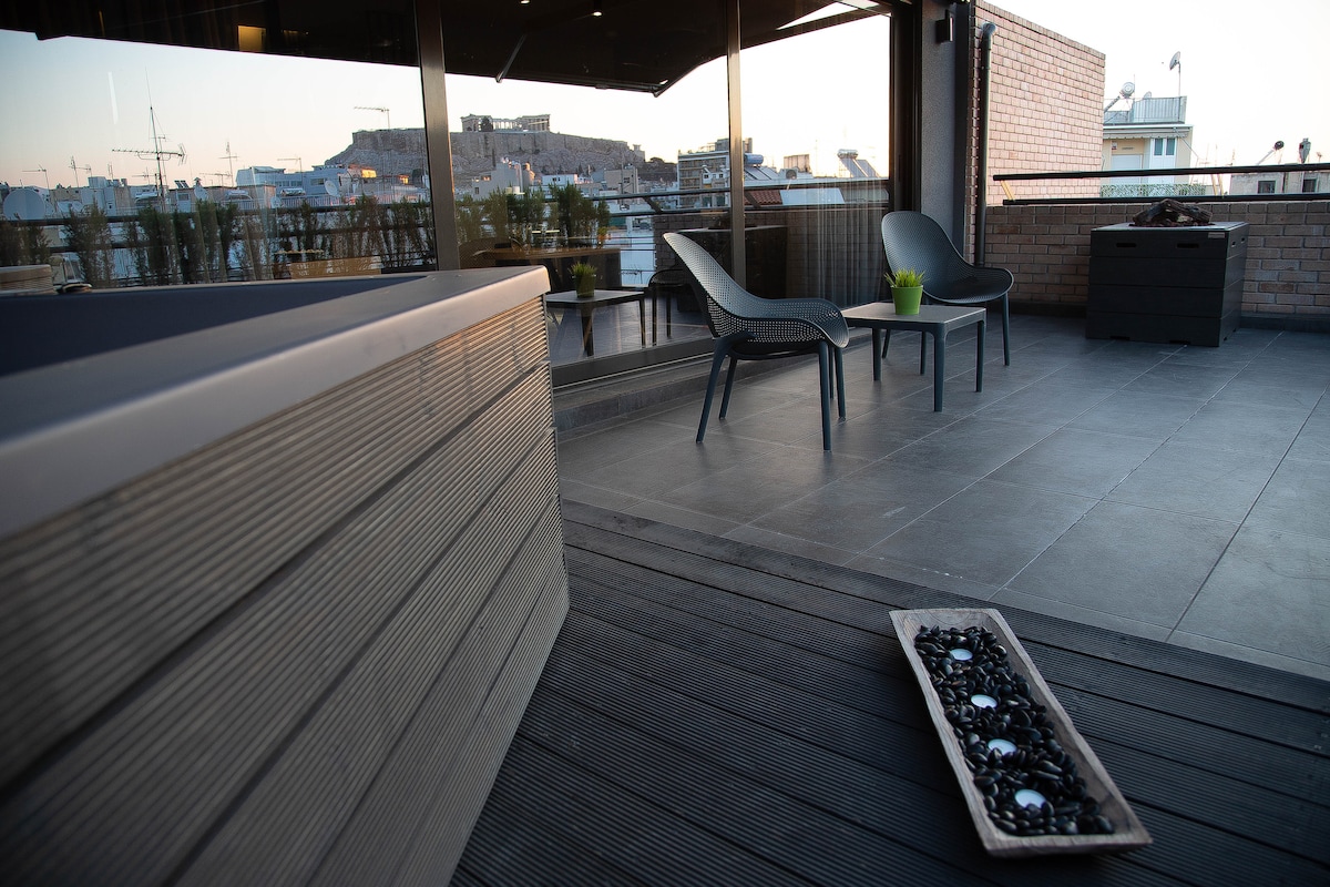 A private terrace features a hot tub and two modern chairs positioned on tiled flooring. Soft lighting highlights a decorative tray with pebbles, while expansive views of the cityscape and Acropolis can be observed in the background.