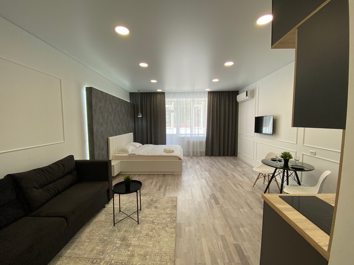 A spacious studio is presented, featuring a king-size bed illuminated by natural light from the large window. A comfortable sofa sits opposite a dining area with a table and chairs. Light-colored flooring enhances the modern design, providing an open and welcoming atmosphere.