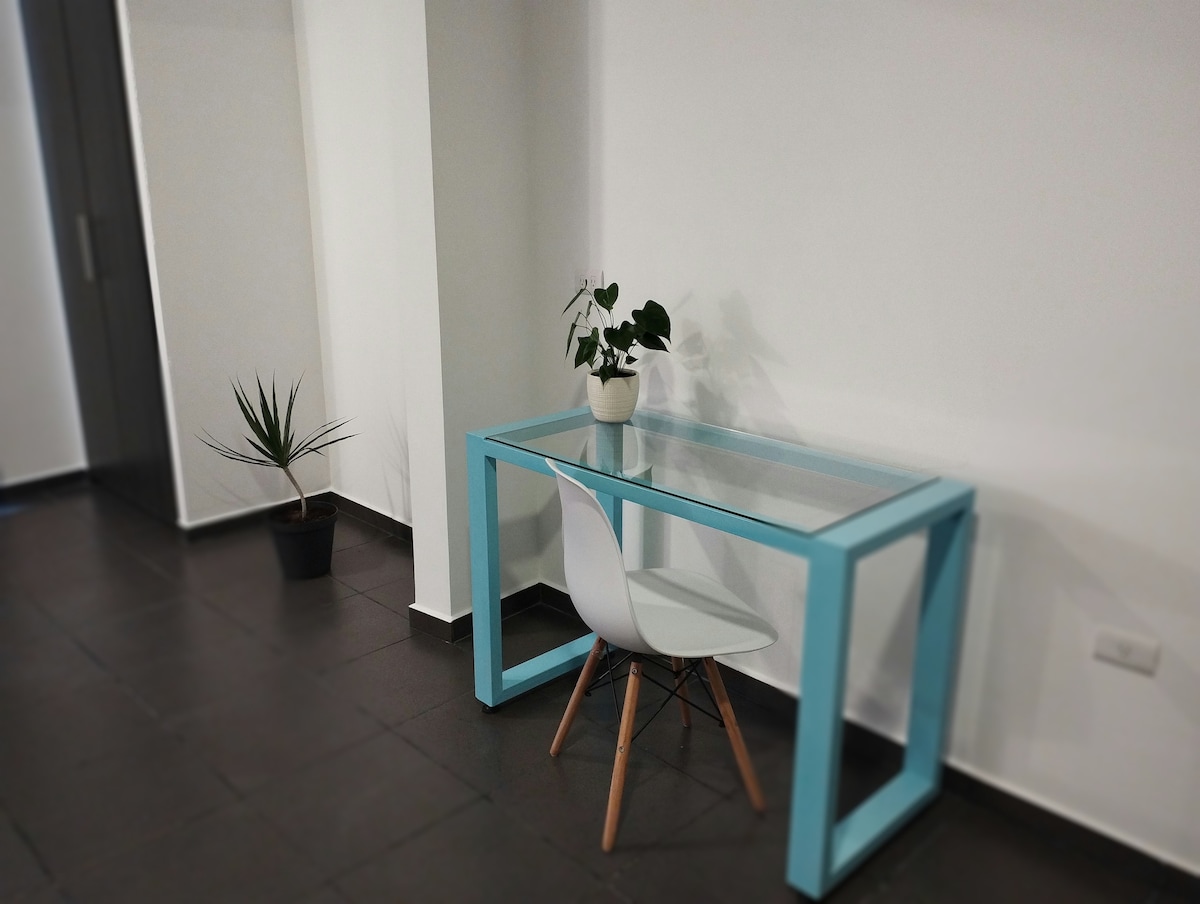 A modern desk with a glass top is positioned against a white wall, accompanied by a simple white chair. A small potted plant sits on the desk, while another tall plant is placed nearby in a dark pot, adding natural elements to the space.