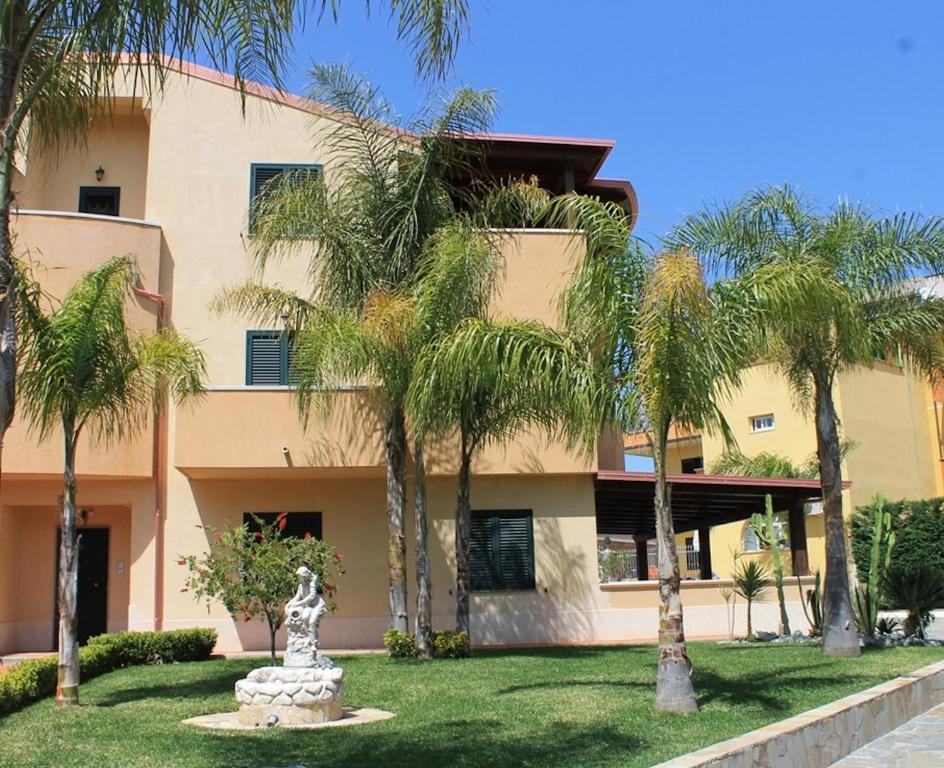 The exterior of the villa is characterized by a vibrant garden with several palm trees and manicured greenery. A decorative fountain is centrally located on the lawn, complementing the entrance of the spacious, two-story structure with large windows.