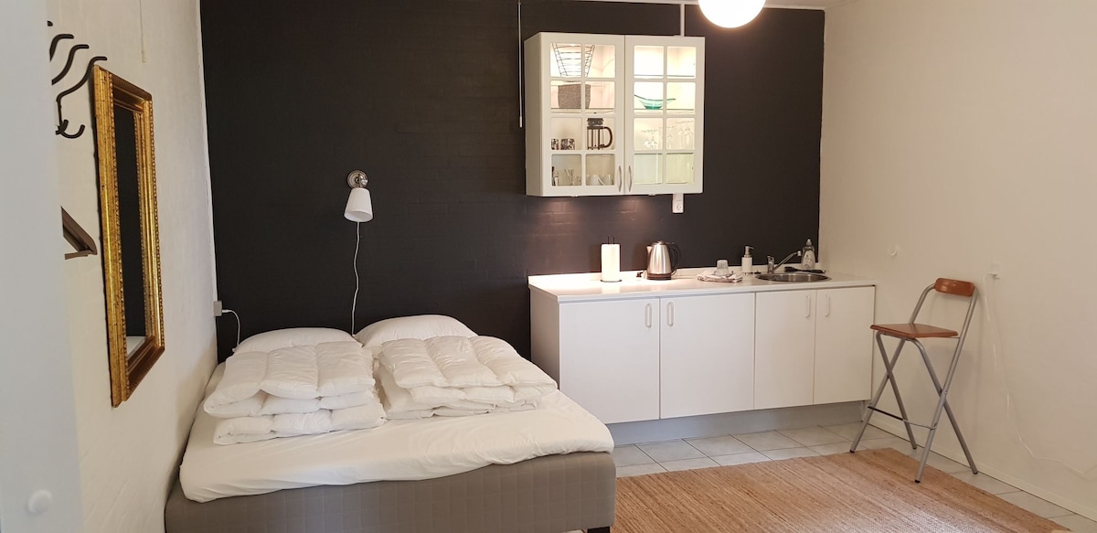 A cozy sleeping area features a spacious bed with folded white bedding. A compact kitchenette is visible against a dark wall, equipped with a kettle and storage. A wooden chair and a woven rug add warmth to the room, creating a comfortable and functional space.