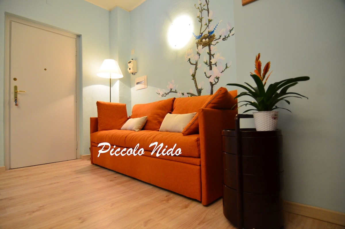 A welcoming lounge area showcases an orange sofa accompanied by accent pillows. A floor lamp provides soft lighting, casting a warm glow. A small potted plant rests beside a wooden side table, with a mural of blossoms enhancing the calming atmosphere.