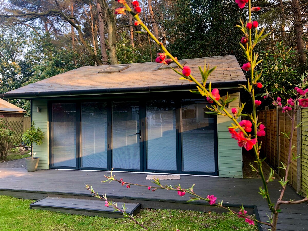 The garden room features a large deck area with a smooth surface leading to expansive glass doors. These doors provide a view into the interior space while being framed by vibrant flowering branches. Surrounding trees contribute to a private, tranquil environment.