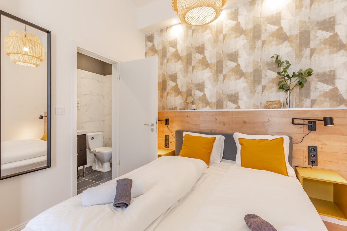 A stylish bedroom is featured with a large bed adorned in crisp white linens and accented with yellow pillows. Natural light is welcomed through a mirror reflecting the space. A neutral-toned bathroom is visible through an open door, showcasing modern fixtures.