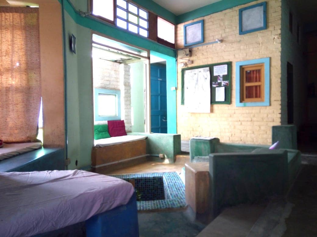 A bright common area features multiple seating areas with green and blue accents. Sunlight filters in through large windows and a door leading outside. A small, tiled pool is visible in the center, surrounded by low seating, creating an inviting space for relaxation and socializing.