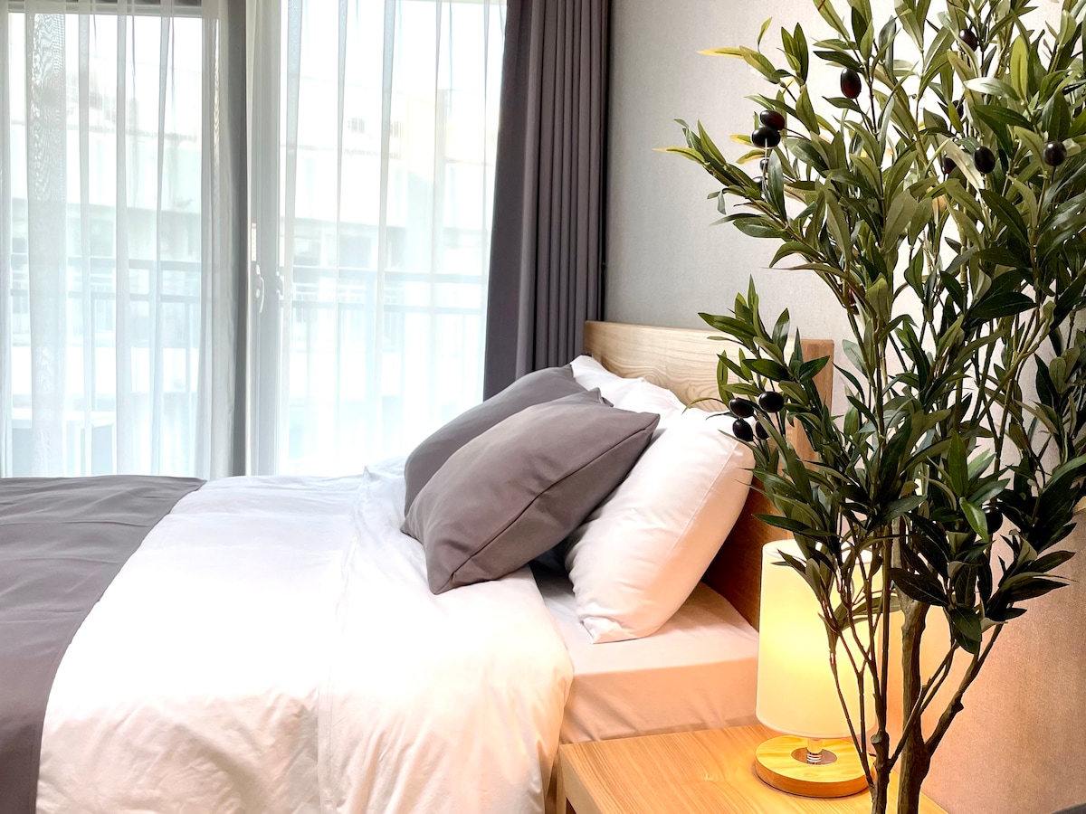 A spacious bed is dressed in crisp white linens and accentuated with gray pillows. A bedside table with a soft lamp is positioned next to the bed, while a decorative leafy plant adds a touch of greenery. Natural light filters through sheer curtains.