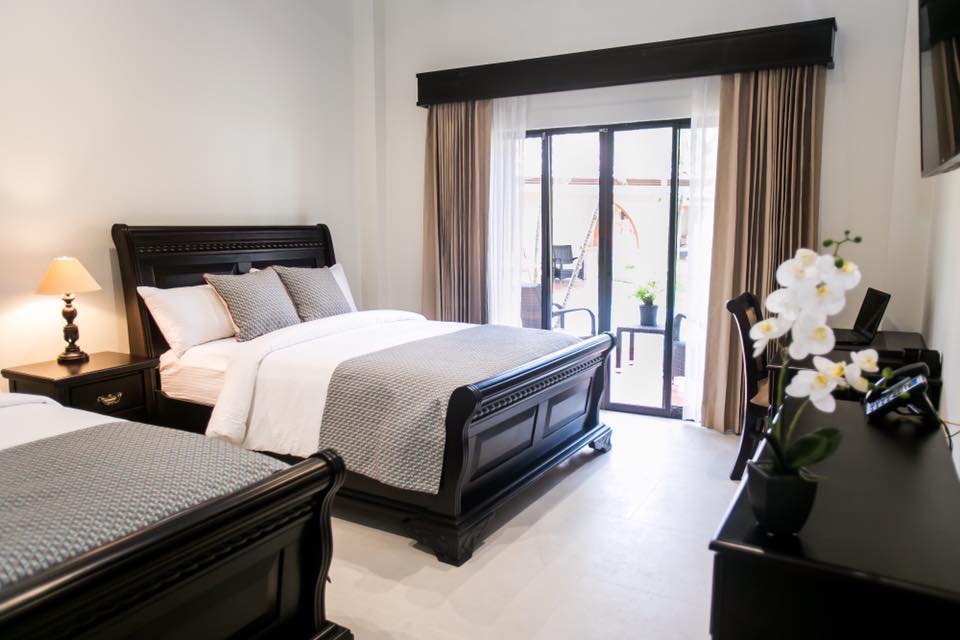 A bright sleeping area features two spacious beds with elegant dark wood headboards. Natural light enters through sliding glass doors that lead to a private patio. A wooden desk and a bedside lamp enhance the minimalist decor, while a potted orchid adds a touch of nature.