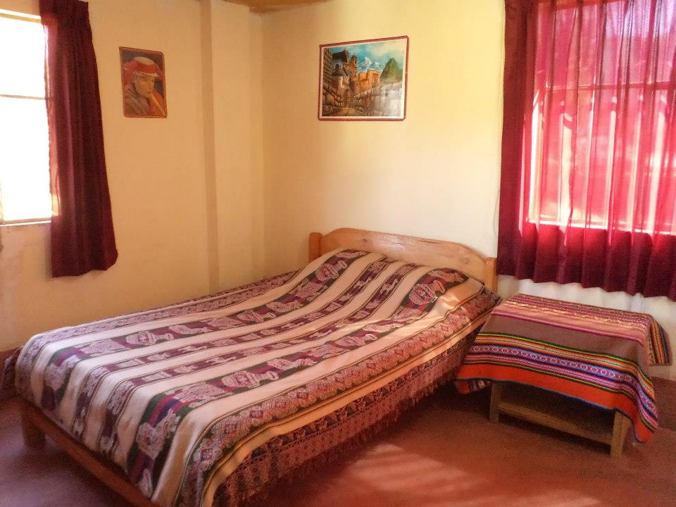 A cozy guest room features a spacious bed adorned with a patterned blanket. A small side table stands beside the bed, and natural light filters through the red curtains. Colorful artwork decorates the walls, adding a warm touch to the inviting space.