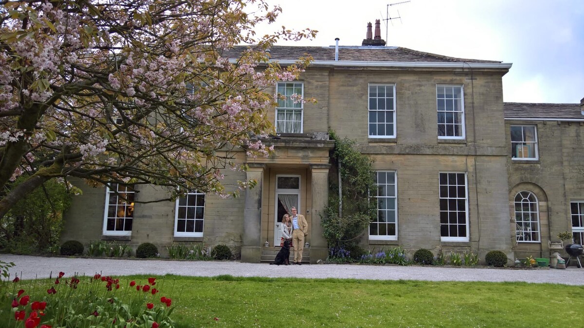 The Georgian house features large windows and a symmetrical façade, framed by a blooming tree in the foreground. A well-maintained pathway leads to the entrance, which is adorned with elegant stonework. Colorful flower beds add vibrancy to the inviting exterior.