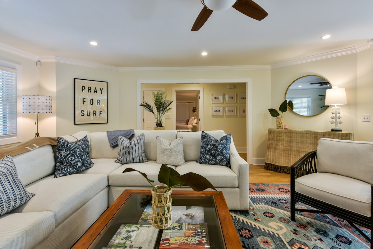 A spacious living room features a large L-shaped sectional sofa adorned with decorative pillows. A coffee table with a glass top holds a vase with greenery. A round mirror and wall art enhance the bright, inviting atmosphere of the room.