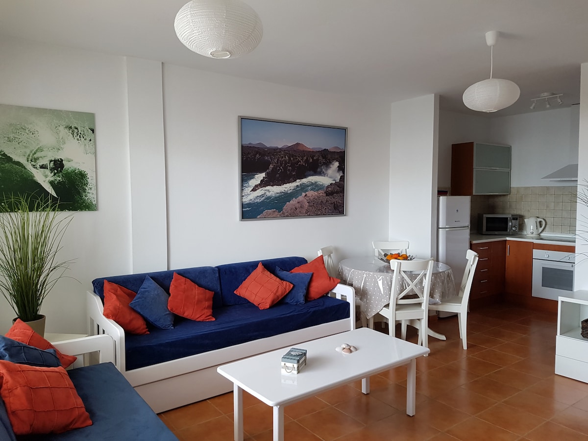 A modern living space features a blue sofa accented with orange cushions, accompanied by a white coffee table. A dining area with a round table and four chairs is present. The kitchen is visible, showcasing wooden cabinetry and modern appliances. Artwork of ocean scenery decorates the wall.