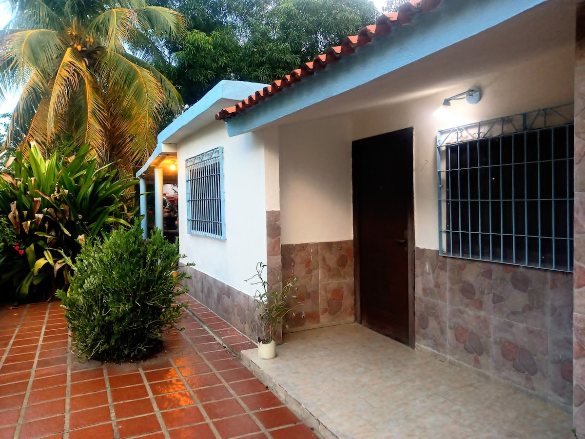 The entrance of the vacation home is framed by lush tropical plants and features a stone walkway paved with red tiles. The building showcases a clean white exterior with a blue roof and barred windows, providing a welcoming and functional appearance.