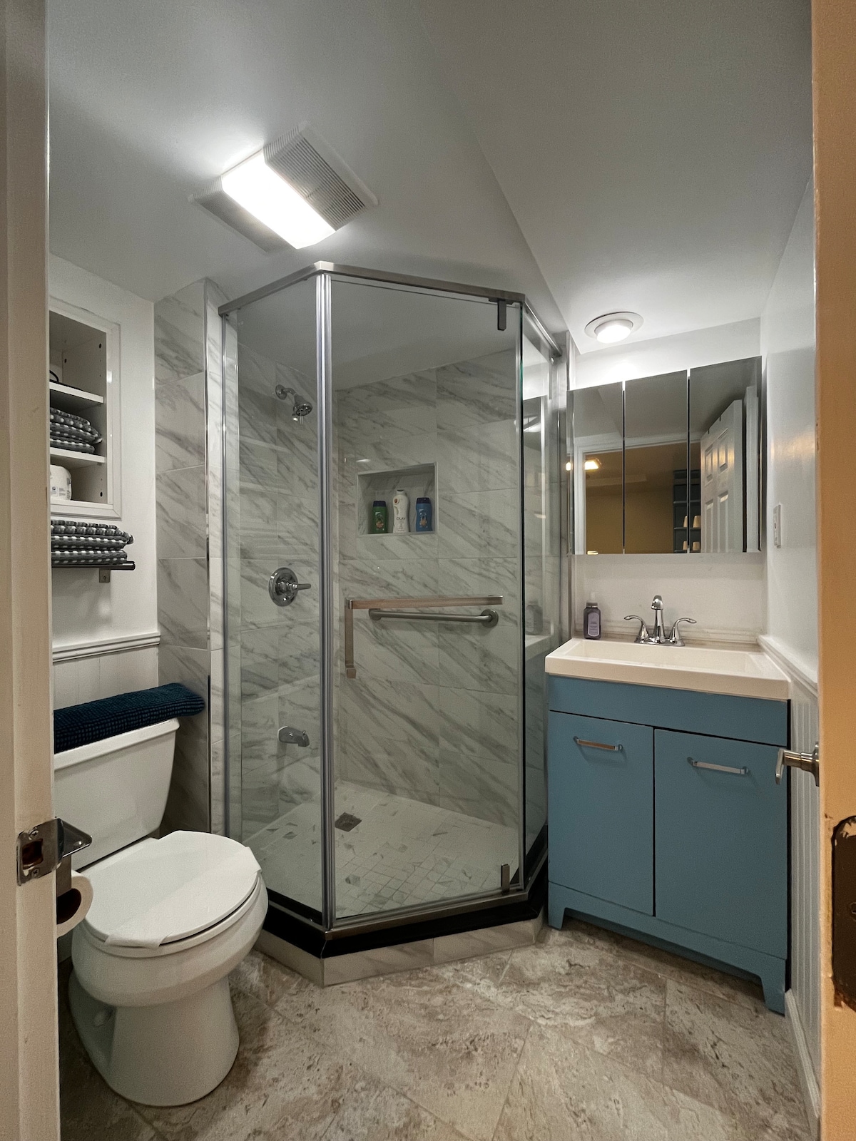 The private bathroom features a glass-enclosed shower with marble walls and a modern sink under a large mirror. A toilet is positioned beside the shower, and shelves above hold neatly arranged toiletries. Soft lighting enhances the functionality of the space.