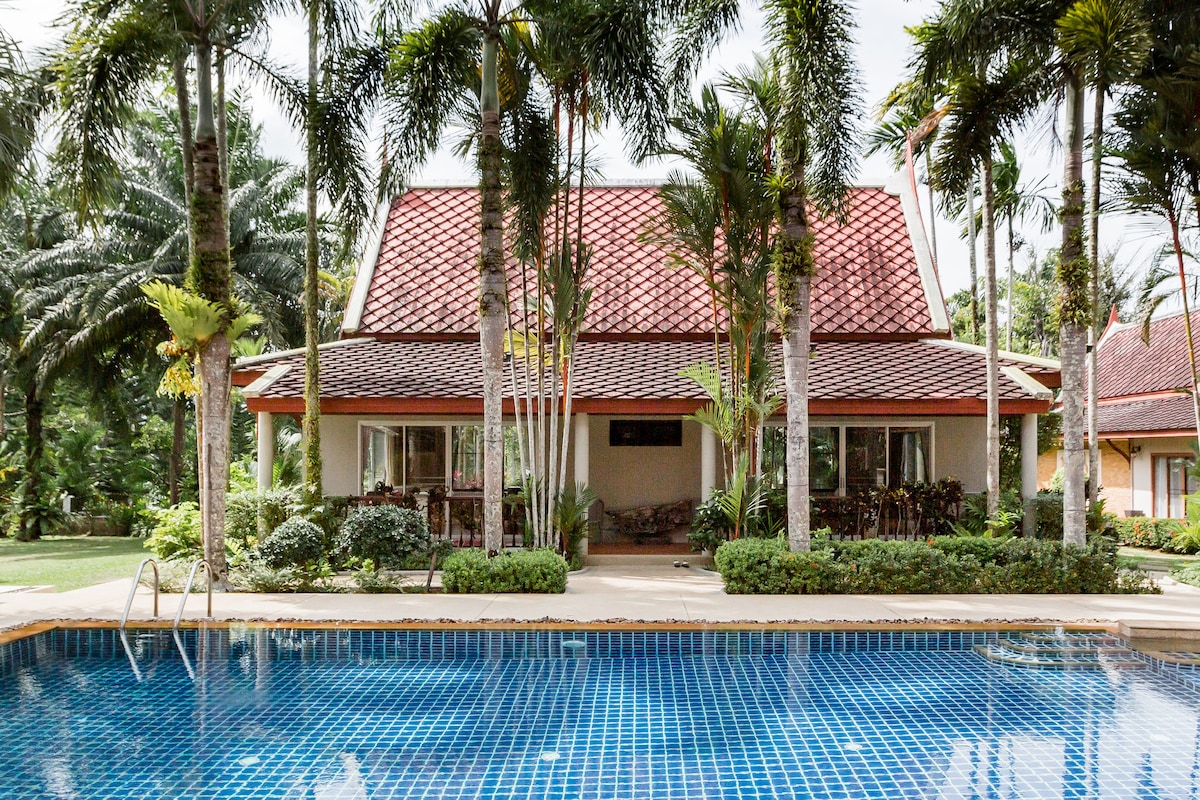 A spacious villa with a traditional Thai design is set against a backdrop of lush palm trees. The swimming pool reflects the vibrant blue tiles, and large windows allow natural light to illuminate the inviting indoor spaces.