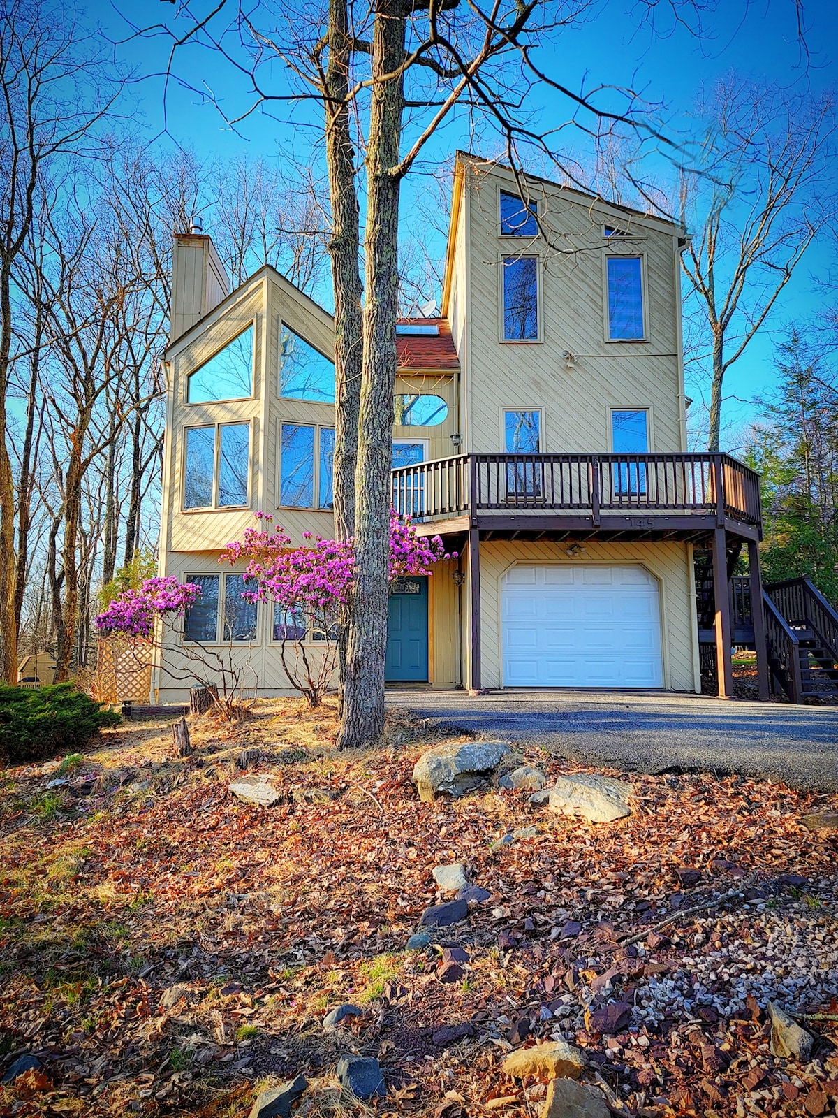 The chalet is designed with a unique, contemporary style, featuring large windows that allow natural light to fill the interior. A deck extends from the upper level, and vibrant purple flowers are visible next to the entrance, surrounded by a landscape of gravel and native vegetation.