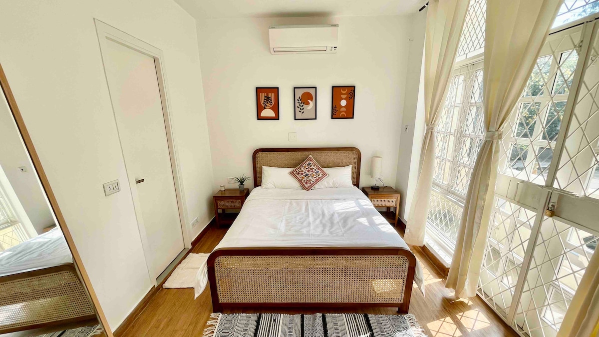 A bright bedroom showcases a comfortable queen-sized bed with a textured rattan headboard. Natural light streams in through large windows adorned with sheer curtains. A small bedside table complements the décor, while artwork featuring faces adds a touch of character to the walls.