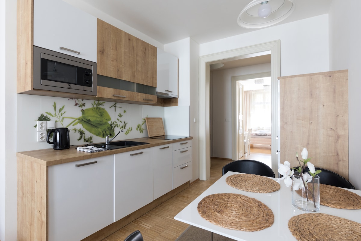 A modern kitchen area features wooden cabinetry paired with white fronts. A glass table is set with woven placemats, complemented by a floral centerpiece. Kitchen appliances, including a microwave and coffee maker, are visible, creating a functional and welcoming atmosphere.