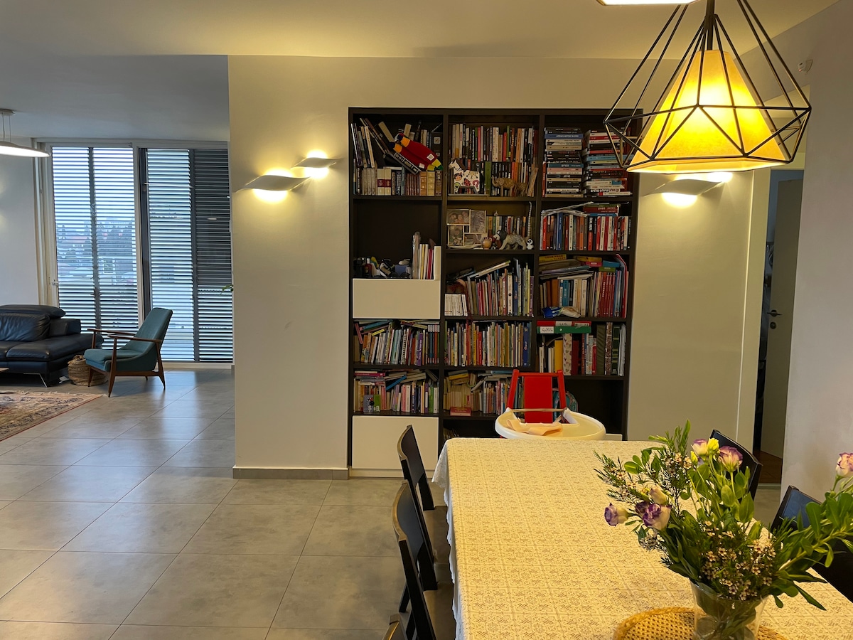 An open living area features a large dining table with a light-colored tablecloth, surrounded by dark chairs. A tall bookshelf filled with books stands against the wall, and a soft illumination from a pendant light highlights the space. Large windows allow natural light to fill the room.