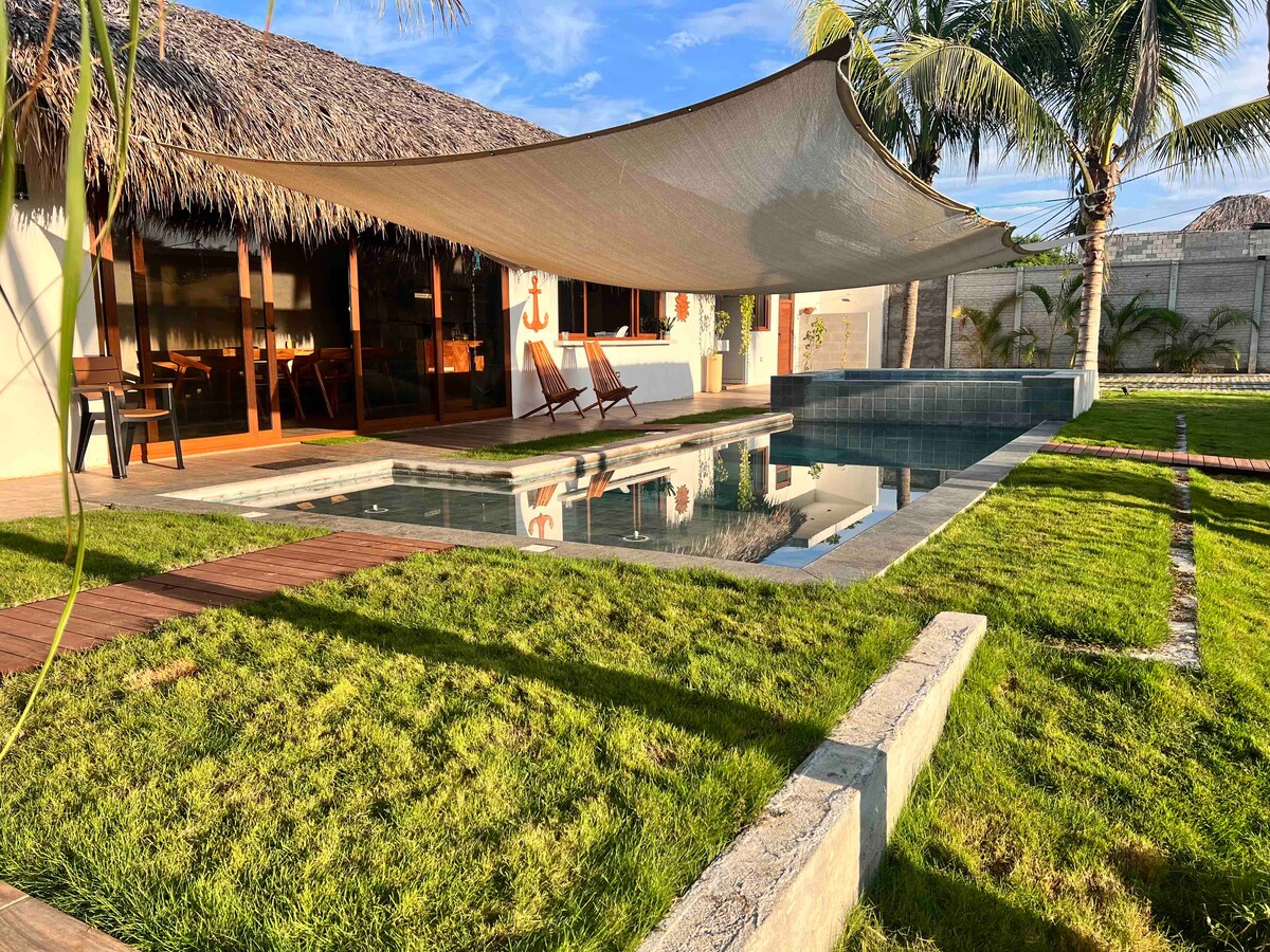 A tranquil outdoor area is presented, featuring a refreshing swimming pool surrounded by lush green grass. In the background, a shaded seating area with two lounge chairs offers a relaxing spot. Nearby, palm trees provide a natural touch to the inviting landscape.