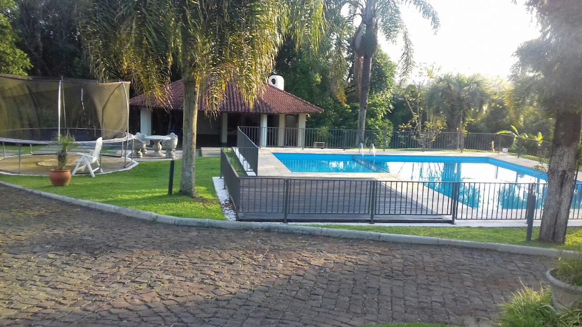 An outdoor area features a sparkling swimming pool surrounded by grass and palm trees. A shaded gazebo is visible in the background, with a trampoline nearby. White lounge chairs are arranged around the pool, providing ample space for relaxation.