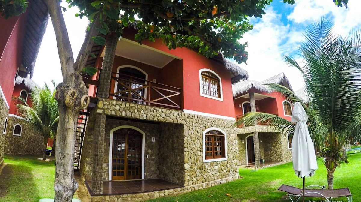 A spacious outdoor area features charming chalets with red walls and traditional thatched roofs. Lush greenery surrounds the grounds, enhancing the tranquil setting. A wooden balcony is visible on the upper level, adding to the inviting character of the architecture.