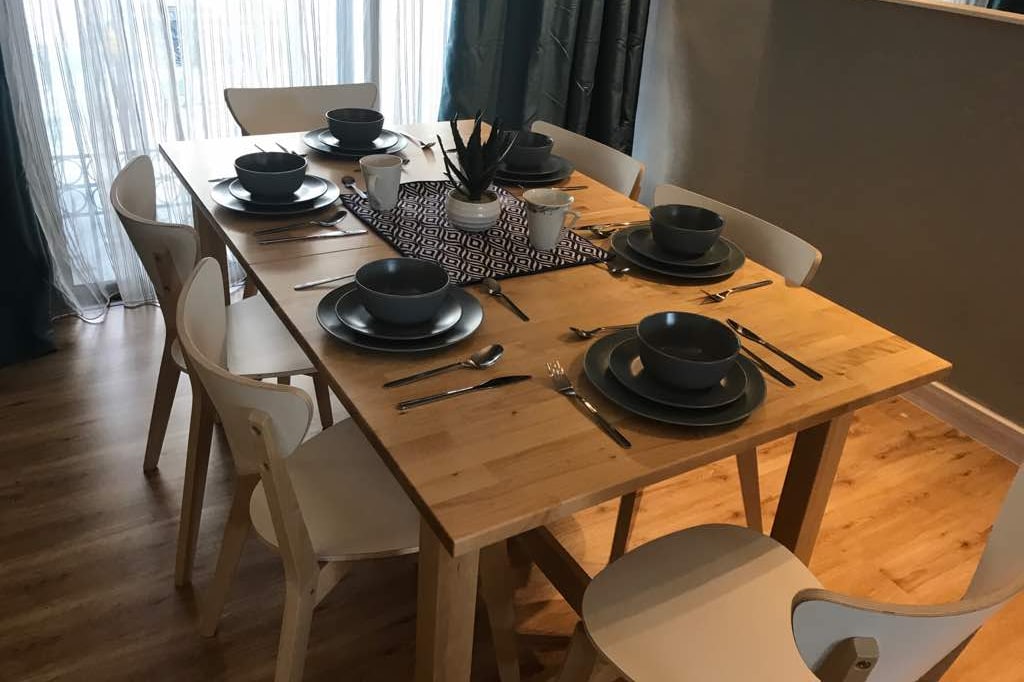 A wooden dining table is set for eight, featuring grey bowls, plates, and cutlery for a meal. Simple table decor includes a patterned runner and a small plant. Light filters through sheer curtains, creating a clean and inviting dining space.