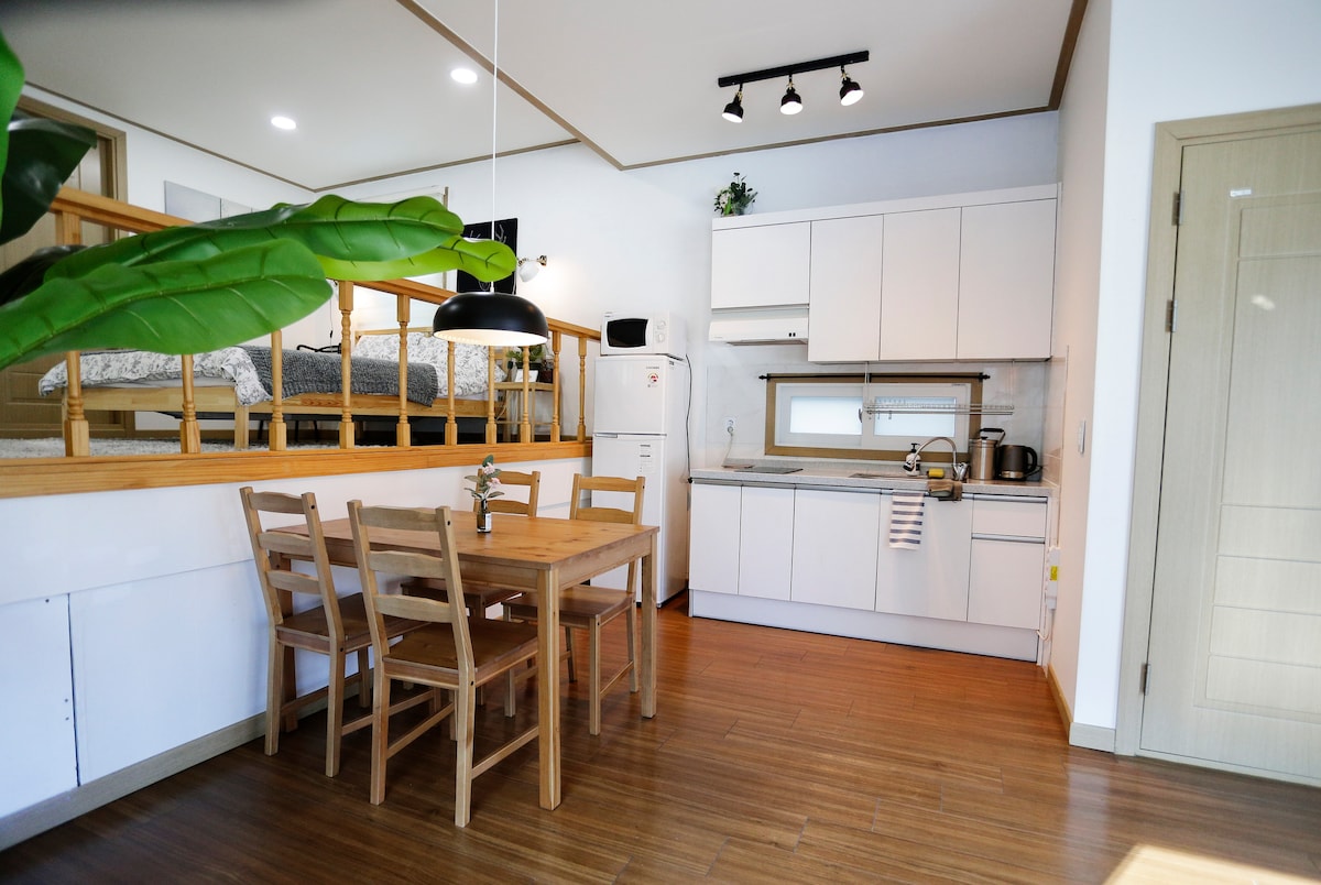 A well-equipped kitchen features modern cabinetry and appliances, including a refrigerator and stove. A wooden dining table, surrounded by four chairs, provides a cozy dining area. A partially visible loft area above adds to the spacious feel of this inviting space.