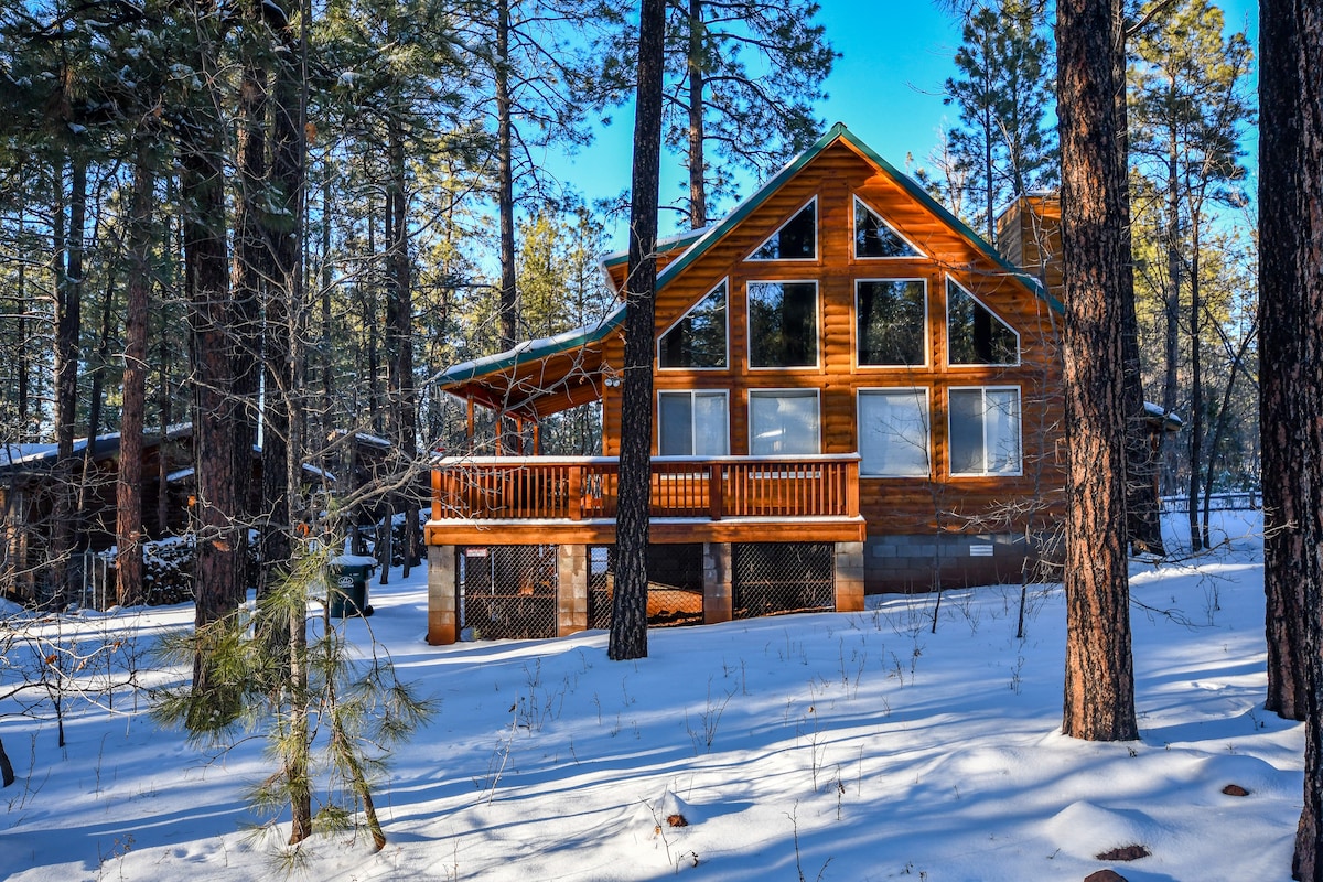The log cabin is nestled among tall trees, featuring large windows that reflect the sunny sky. A spacious wrap-around porch invites relaxation, while a blanket of snow covers the ground, enhancing the serene forest setting.