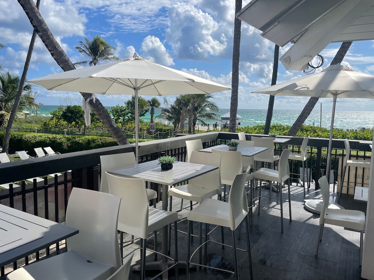 An outdoor dining area is shown with several high-top tables and white chairs. Umbrellas provide shade, while palm trees sway in the background. The ocean glistens under a partly cloudy sky, creating a relaxed atmosphere for enjoying meals by the beach.